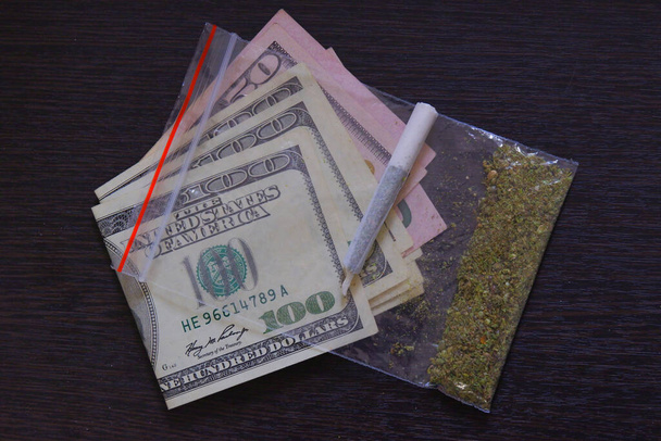 Ένα σακουλάκι μαριχουάνα ή χόρτο σε δολάρια. Έννοια του εμπορίου ναρκωτικών. Κομμένο στέκι κάνναβης σε φόντο καφέ ξύλου από κοντά - Φωτογραφία, εικόνα