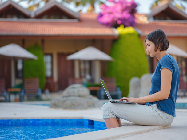 Young business woman working at computer by hotel pool. Young lady downshifter working at laptop and enjoys and relaxed environment, working day. Online freelance work on vacation. - Foto, afbeelding