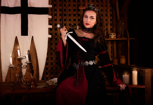 Fantastic medieval woman with a dagger in her hands. Royal velvet vintage red dress with gold, princess, queen, brunette with long hair. Castle, dusk, night, luxury - Фото, изображение