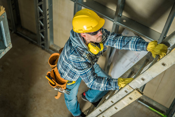 Фото профессионального подрядчика в жёлтой шляпе, поднимающегося по лестнице на строительной площадке. Строительные инструменты и оборудование. - Фото, изображение