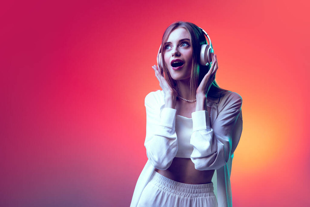 Portrait of young emotive woman posing, listening to music in headphones isolated over red background in neon light. Concept of beauty, lifestyle, youth, emotions, facial expression, ad. - Photo, image