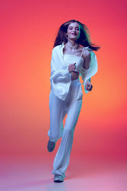 Portrait of young cheerful woman posing in a run isolated over red background in neon light. White clothes. Concept of beauty, lifestyle, youth, emotions, facial expression, ad. - Photo, image