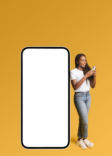 Sorridente ragazza attraente in piedi vicino smartphone enorme con schermo vuoto, sorrisi, hipster donna pubblicità nuova applicazione o sito web, mockup - Foto, immagini