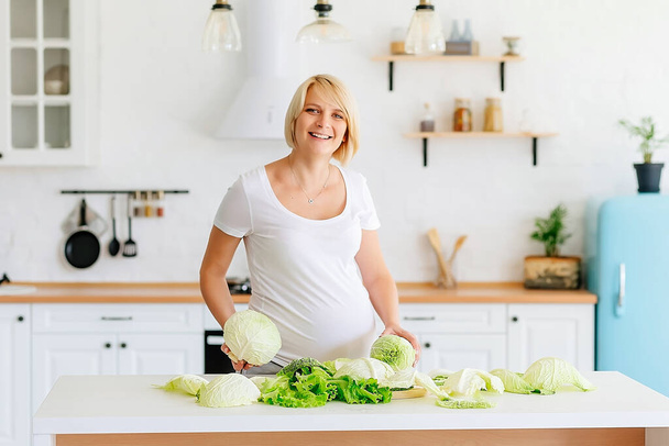 Linda menina loira grávida loira na cozinha em estilo escandinavo na mesa analisa repolho verde. Foto de alta qualidade - Foto, Imagem