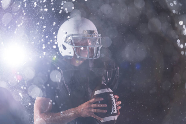 American Football Athlete Warrior Standing on a Field Holds his Helmet and Ready to Play. Player Preparing to Run, Attack and Score Touchdown. Rainy Night with Dramatic lens flare and rain drops. High - Foto, Bild