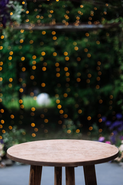 Round wooden table on a green nature with light bulbs background. Outdoor table at desk. Grass, greens and party decoration bushes in the background - Photo, Image