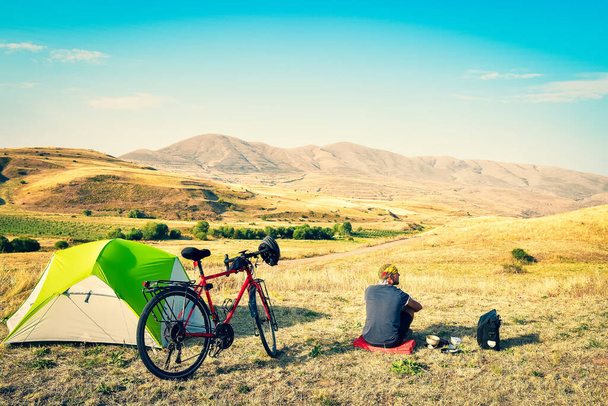 Caucasian man sit by touring bicycle in mountains on side of green tent have break time rest in morning. Solo travel journey. Long-term travel around the world - Photo, Image
