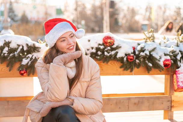 Téli portré fiatal felnőtt gyönyörű nő Mikulás kalap. Karácsonyi hangulat. Havazik. Téli szépség divat koncepció. Csinos fiatal nő ül padon szabadban közel karácsonyi koszorú - Fotó, kép