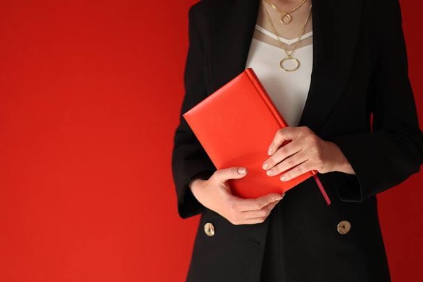 abgeschnittenes Foto einer Geschäftsfrau, die ein Notizbuch auf rotem Hintergrund hält. Erfolgreiche Frau im schwarzen Anzug mit kurzen Haaren blickt in die Kamera. Business, Karriereerfolg, Ausbildungskonzept - Foto, Bild