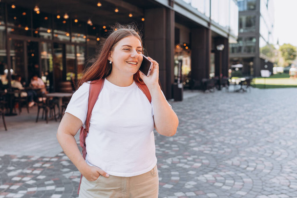Beautiful stylish woman in trendy wear spending time on street using smartphone. Happy Redhead girl talking on the mobile phone, warm sunny day. Urban lifestyle concept - Fotoğraf, Görsel