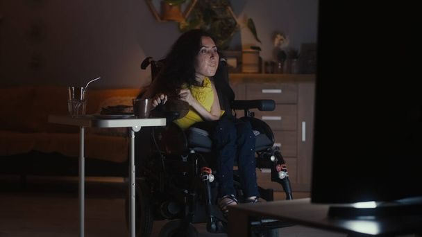 Woman with disability in a modern motorized wheelchair driving into living room and watching TV series in the evening while having dinner - Foto, Bild
