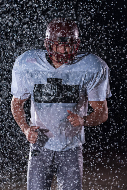 American Football Athlete Warrior Standing on a Field Holds his Helmet and Ready to Play. Player Preparing to Run, Attack and Score Touchdown. Rainy Night with Dramatic lens flare and rain drops. High - Photo, Image