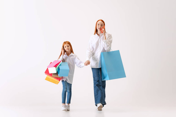Busy ladies. Beautiful young redhaired mother and daughter going shopping with shopping bags isolated on white background. Concept of black Friday, cyber Monday, sales - Фото, изображение