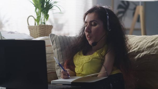 Женщина с ограниченными возможностями в наушниках сидит на диване, записывая в блокнот и разговаривая по видеосвязи во время учебы онлайн на ноутбуке дома - Фото, изображение