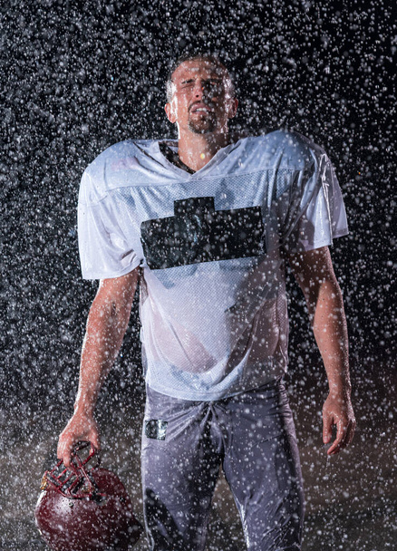 American Football Athlete Warrior Standing on a Field Holds his Helmet and Ready to Play. Player Preparing to Run, Attack and Score Touchdown. Rainy Night with Dramatic lens flare and rain drops. High - Valokuva, kuva