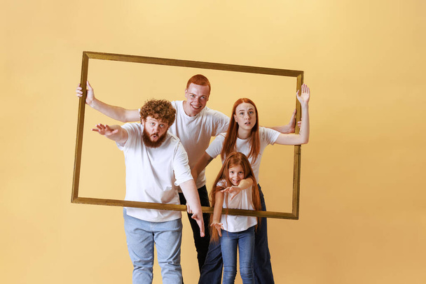 Creative portrait. Joyful redhead family looking through picture wooden frame together. Young men, pretty girl and kid spend time together. Concept of emotions, fun, fashion, relationship - Fotó, kép