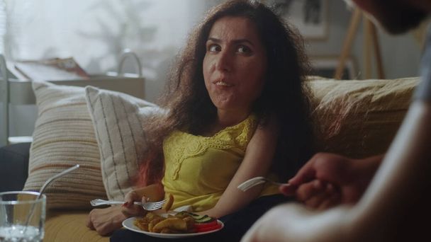 Жена со спинальной мышечной атрофией и муж улыбается и разговаривает во время еды вкусный ужин в гостиной на диване - Фото, изображение
