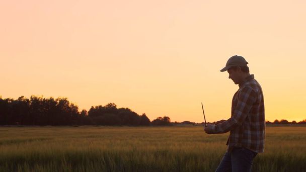 Farmer with a laptop computer in front of a sunset agricultural landscape. Man in a countryside field. The concept of country life, food production, farming and technology. - Photo, image
