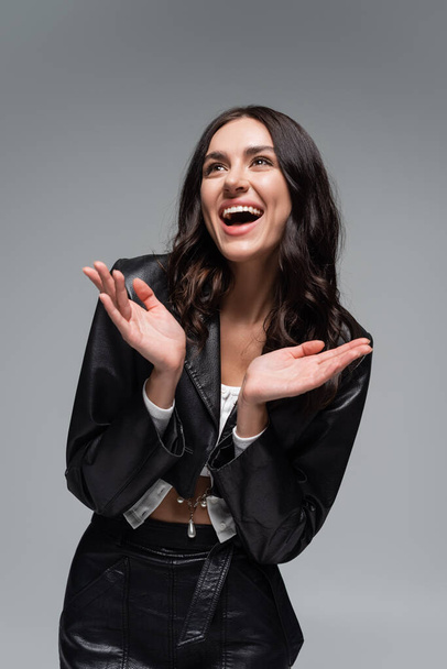 excited young woman in stylish black leather jacket gesturing isolated on grey - 写真・画像