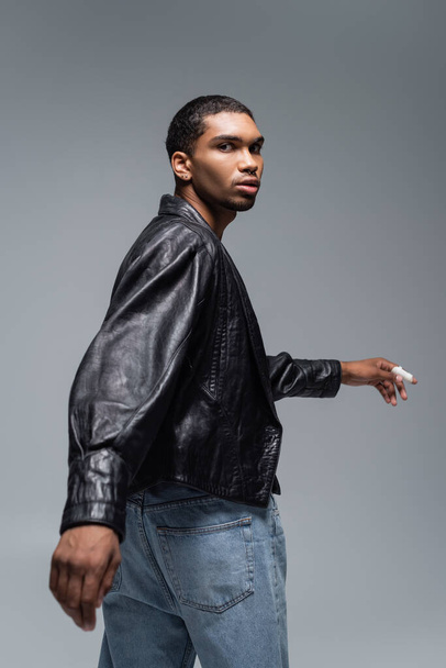 young african american man in jeans and black leather jacket looking at camera isolated on grey  - Fotografie, Obrázek