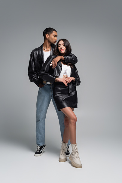 full length of stylish african american man posing with hand in pocket and hugging young woman in leather outfit on grey - Foto, Bild