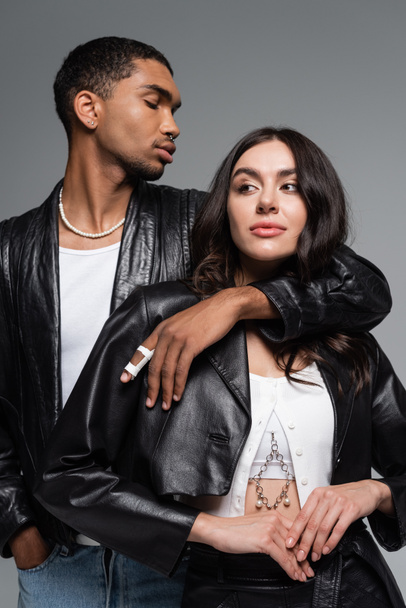 stylish african american man posing with hand in pocket and hugging brunette woman in leather jacket isolated on grey - Foto, immagini