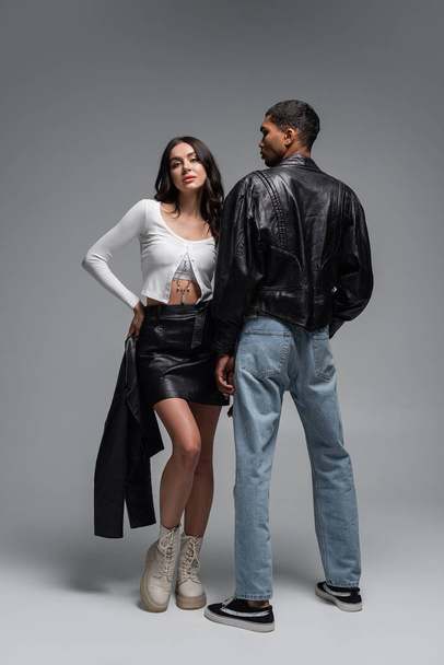 full length of woman in leather skirt holding black jacket near stylish african american man on grey  - Foto, imagen