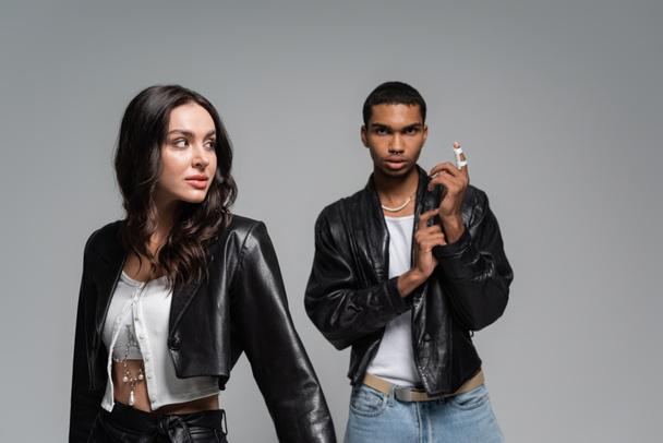 young and stylish multiethnic couple in black leather jackets posing isolated on grey - 写真・画像