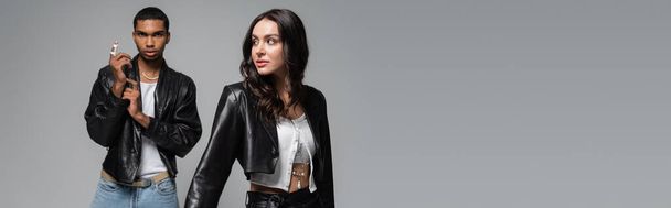 young and stylish multiethnic couple in black leather jackets posing isolated on grey, banner - Photo, Image