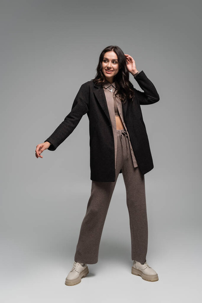 full length of joyful young woman in black blazer and knitted trousers posing on grey - Fotografie, Obrázek