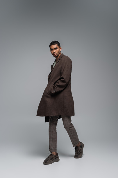 full length of young african american man in autumnal woolen coat looking at camera and posing with hand in pocket on grey  - Fotografie, Obrázek