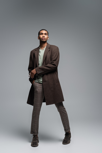 full length of young african american man in autumnal woolen coat looking at camera and standing with hand in pocket on grey  - Foto, Bild