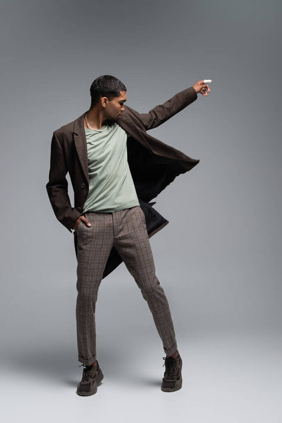 full length of young african american man in autumnal woolen coat posing with hand in pocket on grey  - Photo, image