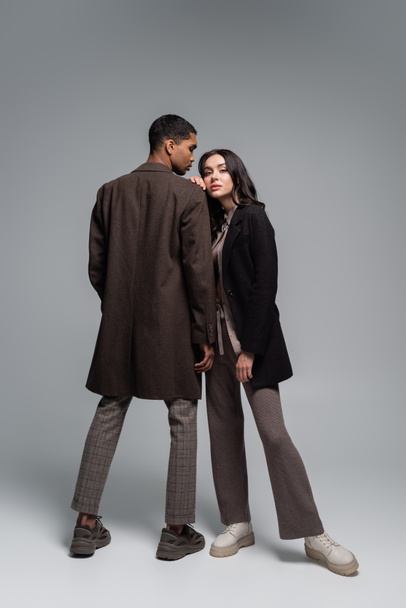 full length of stylish interracial couple in autumnal outfits posing together on grey - Photo, Image