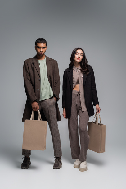 full length of stylish interracial man and woman in autumnal outfits holding shopping bags on grey - Photo, Image