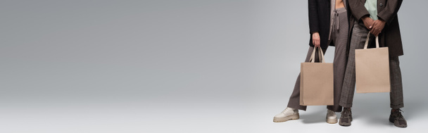 περικοπή άποψη του κομψό διαφυλετικό ζευγάρι σε φθινοπωρινά ρούχα κρατώντας τσάντες ψώνια σε γκρι, πανό - Φωτογραφία, εικόνα