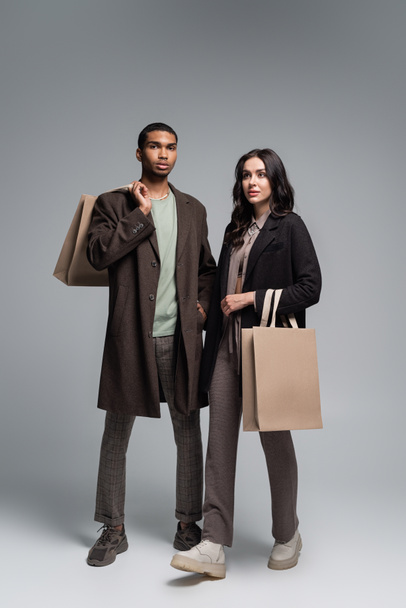 full length of stylish interracial couple in autumnal outfits holding shopping bags on grey - Zdjęcie, obraz