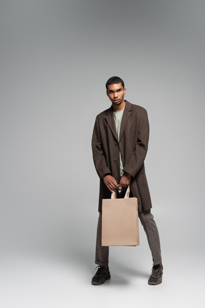 pleine longueur de élégant homme afro-américain en manteau d'automne et baskets posant avec des sacs à provisions sur gris - Photo, image