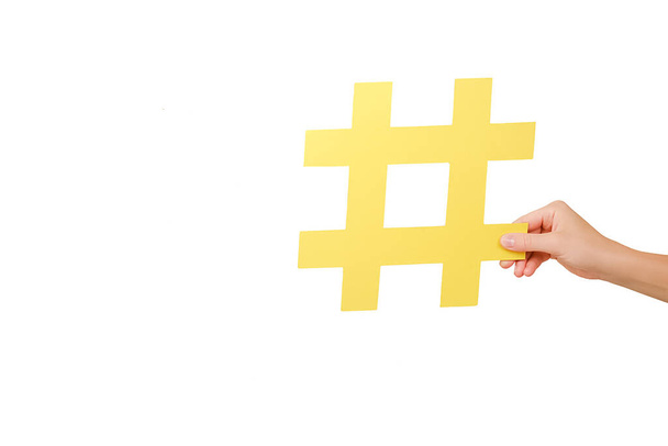 Close up of woman hand holding large yellow paper hashtag symbol, hash sign of famous media content, social media marketing and blog promotion, posing over white studio background wall with copy space - Foto, Imagem