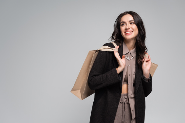 cheerful young woman in black blazer holding shopping bags isolated on grey - Fotó, kép
