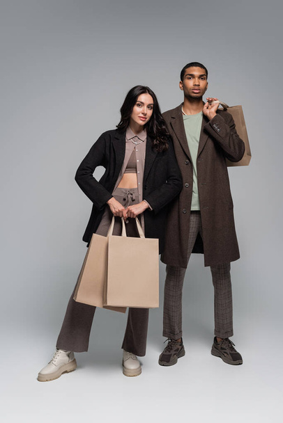 full length of fashionable interracial couple in stylish outfits holding shopping bags on grey - Foto, Imagem