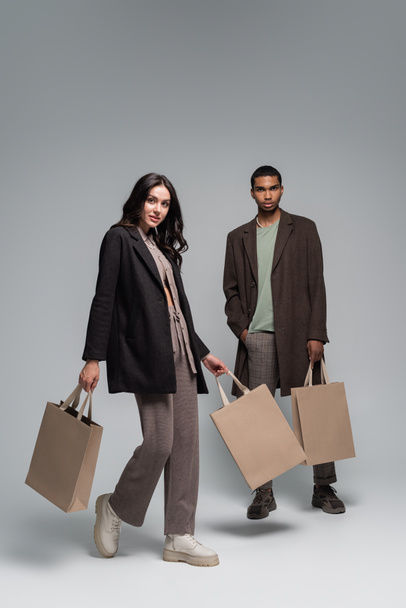 full length of interracial couple in stylish outfits holding shopping bags and walking on grey - Photo, Image