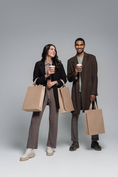 pełna długość szczęśliwy międzyrasowy para w stylowe stroje trzymając torby na zakupy i papierowe kubki na szary - Zdjęcie, obraz