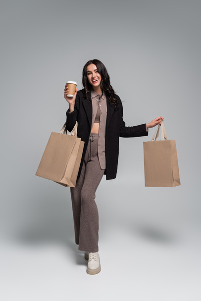 full length of cheerful young woman holding paper cup with coffee to go and shopping bags on grey - Φωτογραφία, εικόνα