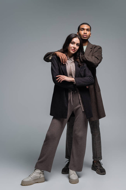 full length of african american man hugging brunette woman in blazer isolated on grey - Foto, afbeelding
