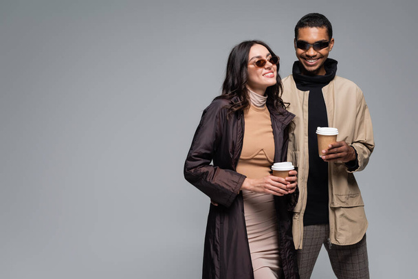 cheerful interracial couple in stylish outfits and sunglasses holding paper cups isolated on grey - Foto, immagini