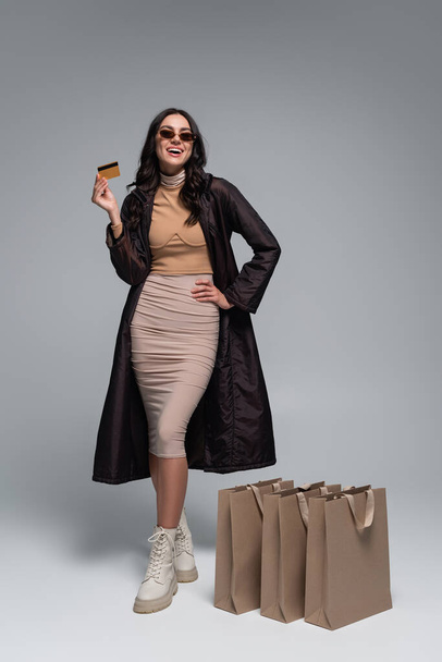 longitud completa de la joven sonriente en traje elegante celebración de la tarjeta de crédito cerca de bolsas de compras en gris - Foto, Imagen