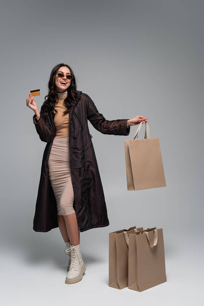 full length of cheerful young woman in stylish outfit holding credit card near shopping bags on grey - Fotografie, Obrázek