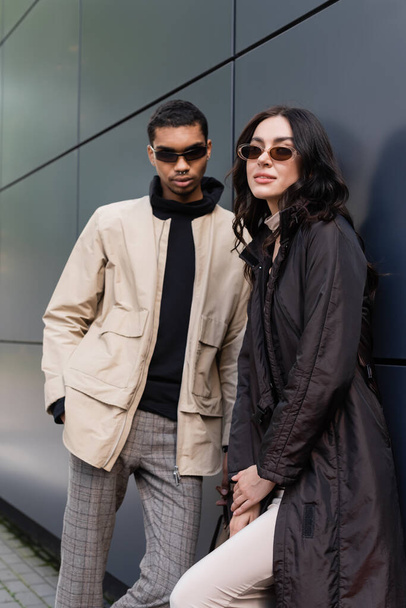 stylish african american man in sunglasses standing with hand in pocket near young woman in coat - Foto, Bild