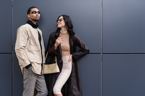 stylish african american man in sunglasses standing with hand in pocket near happy young woman in coat - Foto, imagen
